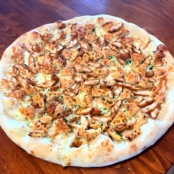 A Mamma Mia's on the Beach pizza with white sauce, chicken, and waffle pieces on a wooden table.